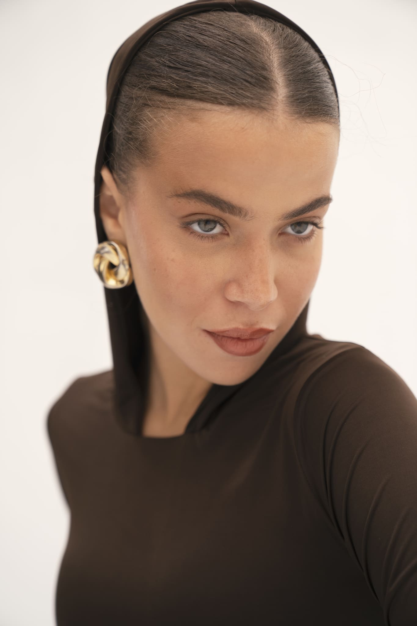 A person with slicked-back hair wearing an Urban Drape Top in brown and a headscarf is looking slightly off-camera. They have striking, light-colored eyes adorned with gold hoop earrings. The softly blurred background focuses attention on their face and expression, highlighting the versatile styling of this contemporary Urban Drape piece.