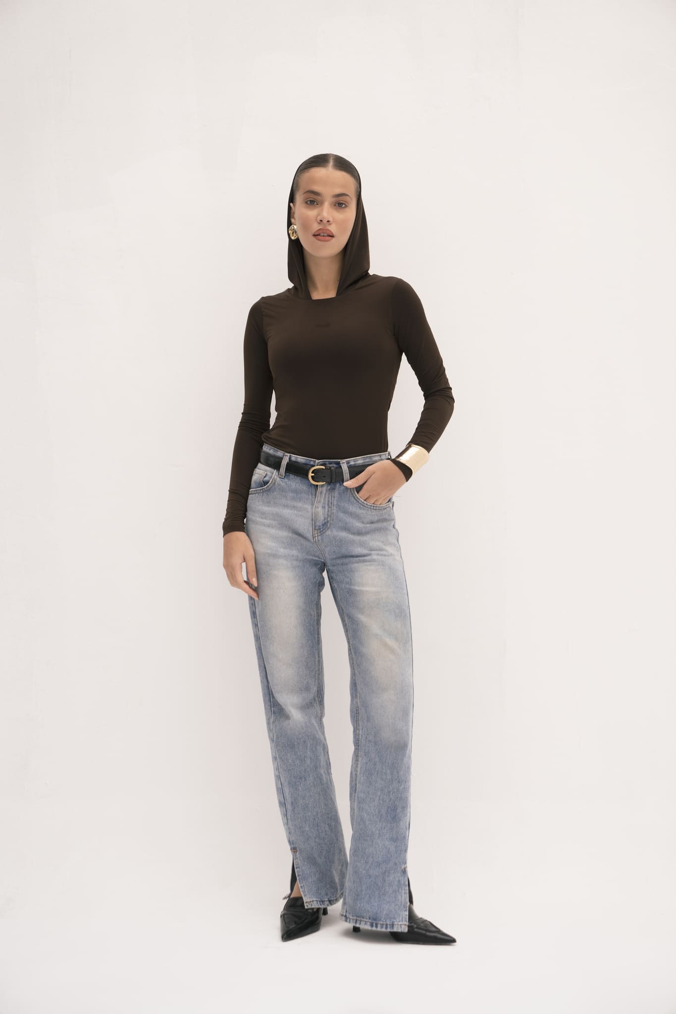 Against a plain white background, a person enhances their modern style with the Urban Drape Top in black, paired with light blue jeans and sleek black pointed shoes. Their long hair cascades elegantly, adorned with gold hoop earrings and accented by a chic belt. Their right hand rests casually in their pocket.