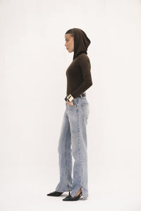 Wearing a black Urban Drape Top, light blue jeans, and black pointed shoes, a person stands in profile against a white background. With hands in pockets, they exude contemporary style and confidence.