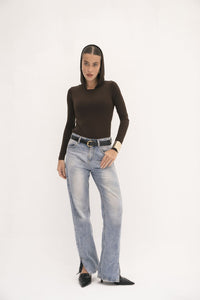 A person poses against a plain white backdrop, demonstrating adaptable styling with the Urban Drape Top paired with light blue jeans and highlighted by a belt. Their straight hair adds to the urban flair of their ensemble, finished off with black shoes. With their left hand in a pocket and their right hand at their side, they exude confidence.