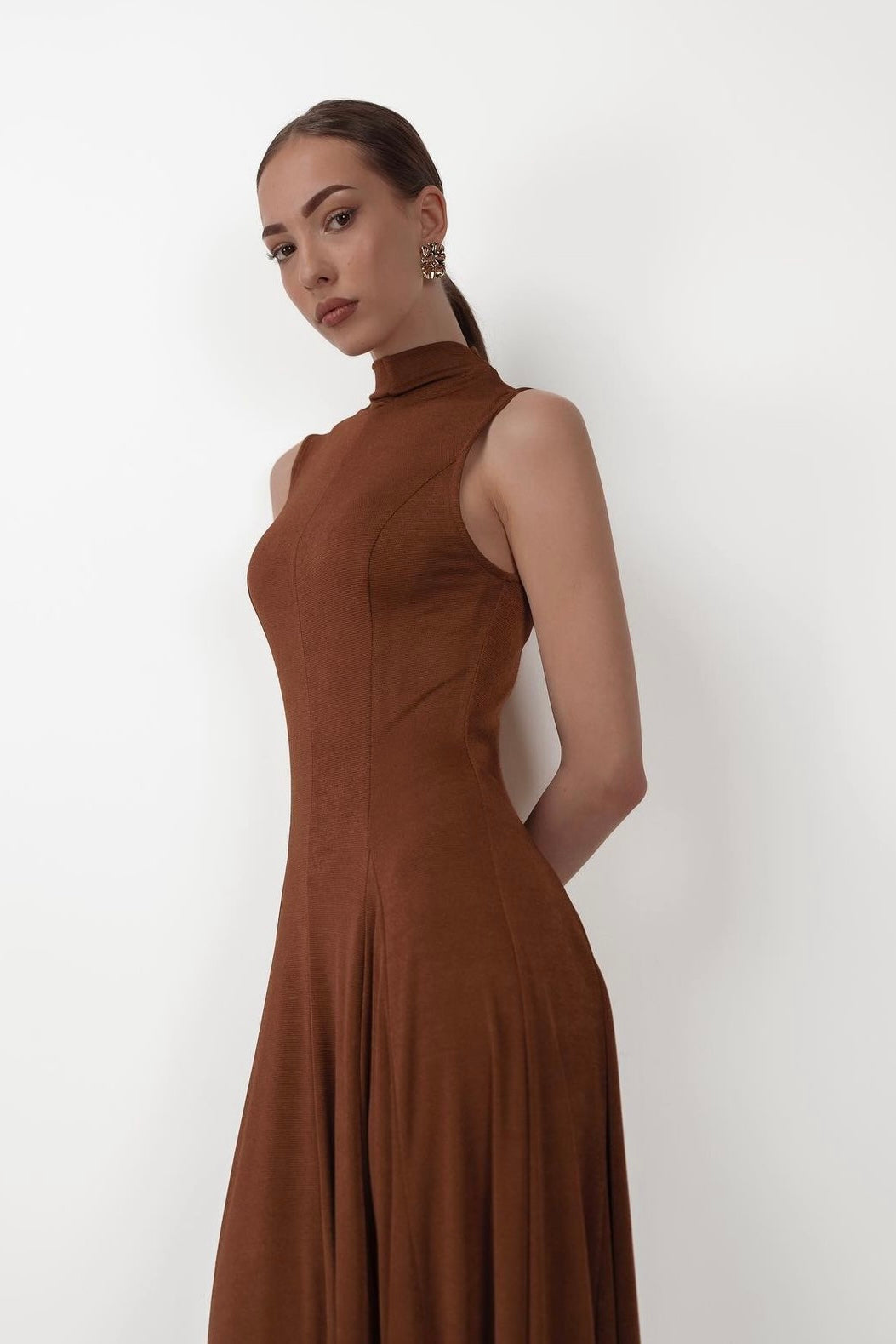 A woman with long brown hair, tied back into a ponytail, is wearing the sleeveless, high-neck Ezra dress. Made of stretchable fabric, she stands against a plain white background with her hands behind her back and looks directly at the camera with a neutral expression.