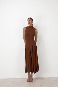 Against a plain white backdrop, a woman poses in an ankle-length brown Ezra dress made from stretchable fabric with pleats. She stands with her arms gently crossed behind her back, glancing slightly to the side. Her sleeveless maxi dress pairs perfectly with open-toed shoes.