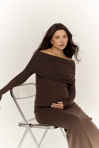 A woman with long dark hair is elegantly seated on a clear chair, dressed in the Madeline knit set. Her off-shoulder top accentuates her form as she lightly touches her belly with one hand, while the other rests gracefully on the chair. She gazes directly at the camera with a neutral expression against a plain white background, highlighting her timeless elegance.