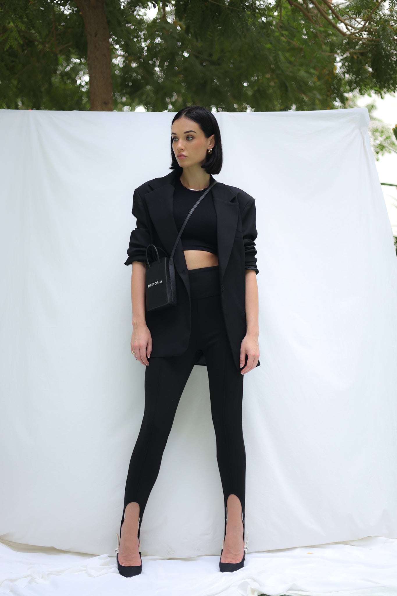 A woman clad in a black crop top, blazer, and Astra Leggings stands against a white backdrop adorned with greenery above. Her leggings feature slits at the ankles, accentuating her black high heels. She holds a small black bag and poses with a neutral expression in this monochromatic outfit.