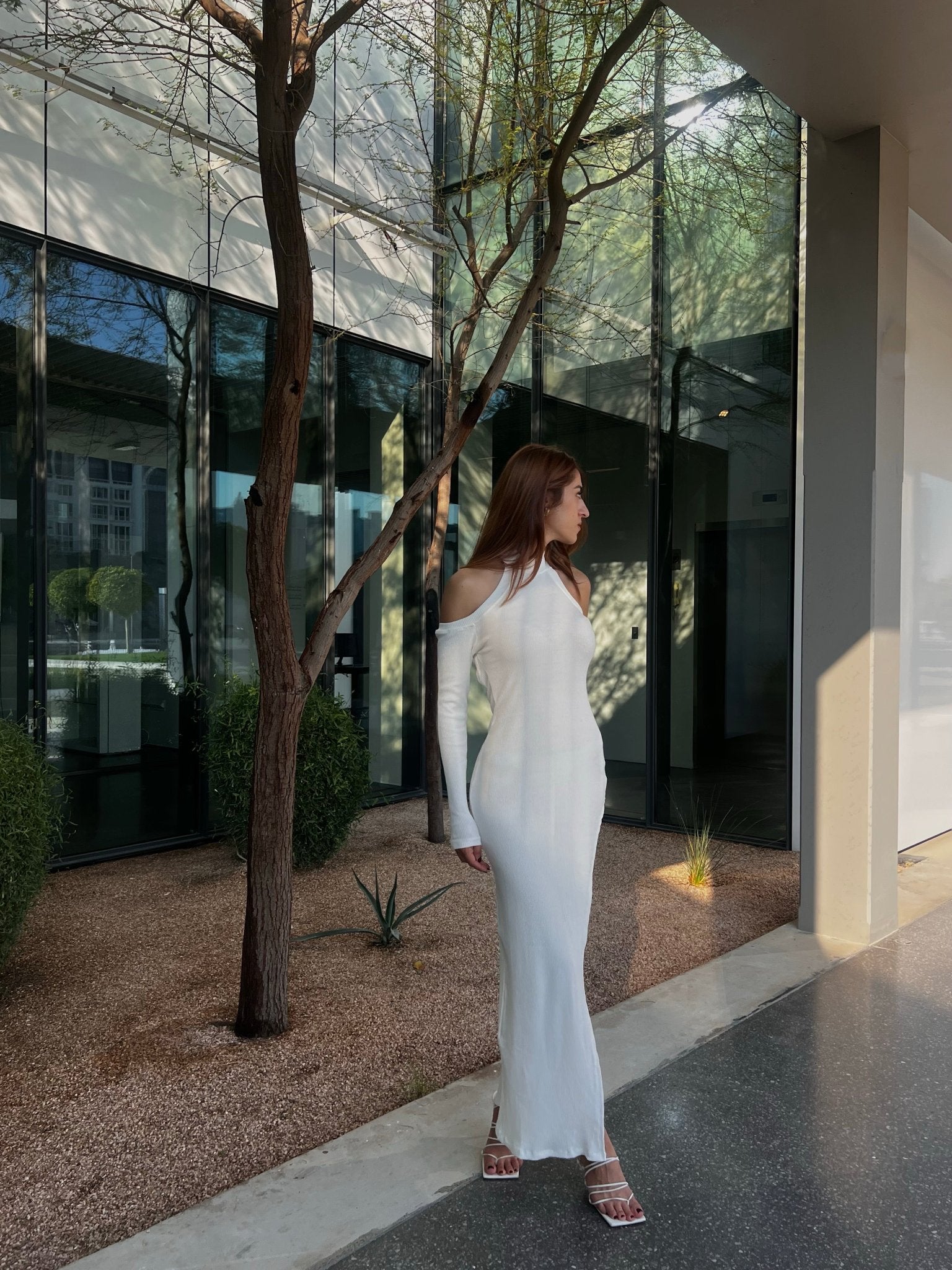 A woman in a sleek Alara dress with cut-out shoulders stands outside a modern building with large glass windows, gazing to the side as her brown hair cascades down her shoulders. The ground is covered with gravel and sparse tall grass, while soft sunlight casts shadows over the scene.