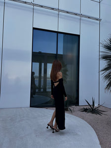 A woman with long brown hair, donning a black Alara dress that features cut-out shoulders and high heels, stands with her back to the camera. She is near a modern building characterized by large glass windows and minimalistic architecture. Nearby, some desert plants are visible.