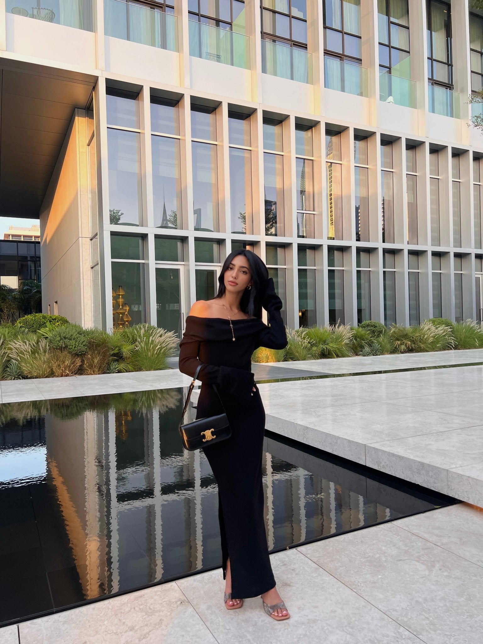 A woman in an off-the-shoulder Abelia knit dress with a side slit stands by a reflective water feature in front of a modern building with large windows. She holds a black handbag and poses with one hand touching her hair. The setting appears to be an urban, contemporary outdoor space.