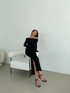 A woman in an Abelia knit dress featuring a black off-the-shoulder design and a thigh-high side slit sits gracefully on the arm of a white modern chair. She complements her dress with black high-heeled sandals and gazes to the side with a neutral expression. The minimalist setting is enhanced by the plain white background.