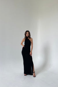 A woman wearing an Aimée dress featuring teardrop cut-outs and an open back adorned with pearls stands confidently in a minimalistic white room. She has long hair, poses with one hand on her hip and the other relaxed by her side, and wears black heels.