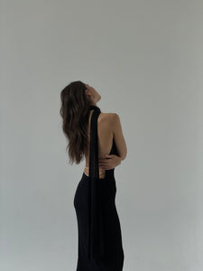 A woman with long, wavy brown hair stands against a plain, light-colored background. She is wearing the Aimée dress, a maxi piece featuring a halter neck and an open back adorned with pearls. Her head is tilted back, and one arm rests on her opposite shoulder.