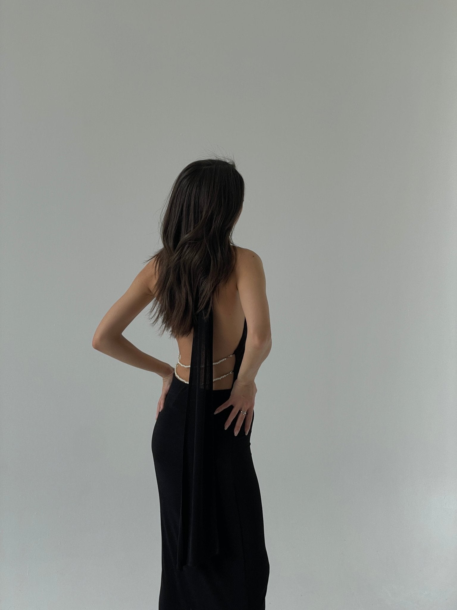 A woman with long, wavy hair stands with her back to the camera, wearing an Aimée dress that features a low open back adorned with pearls and thin straps. She places one hand on her hip while looking to her side against a plain, light-colored background.