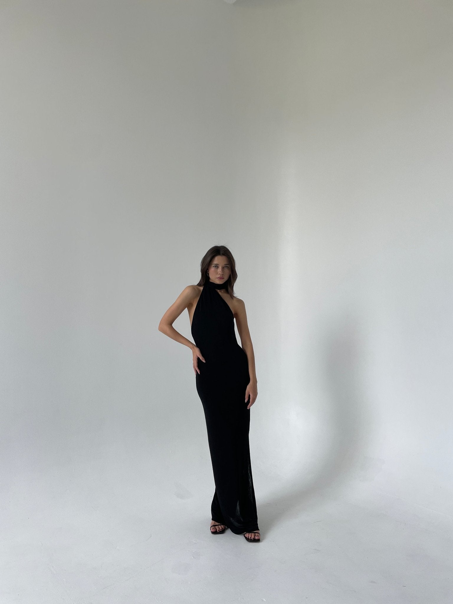 A woman stands in a minimalist white studio, wearing an elegant Aimée dress with a halter neckline and teardrop cut-outs. She has one hand on her hip and looks directly at the camera. The setting is simple, emphasizing her and the attire.