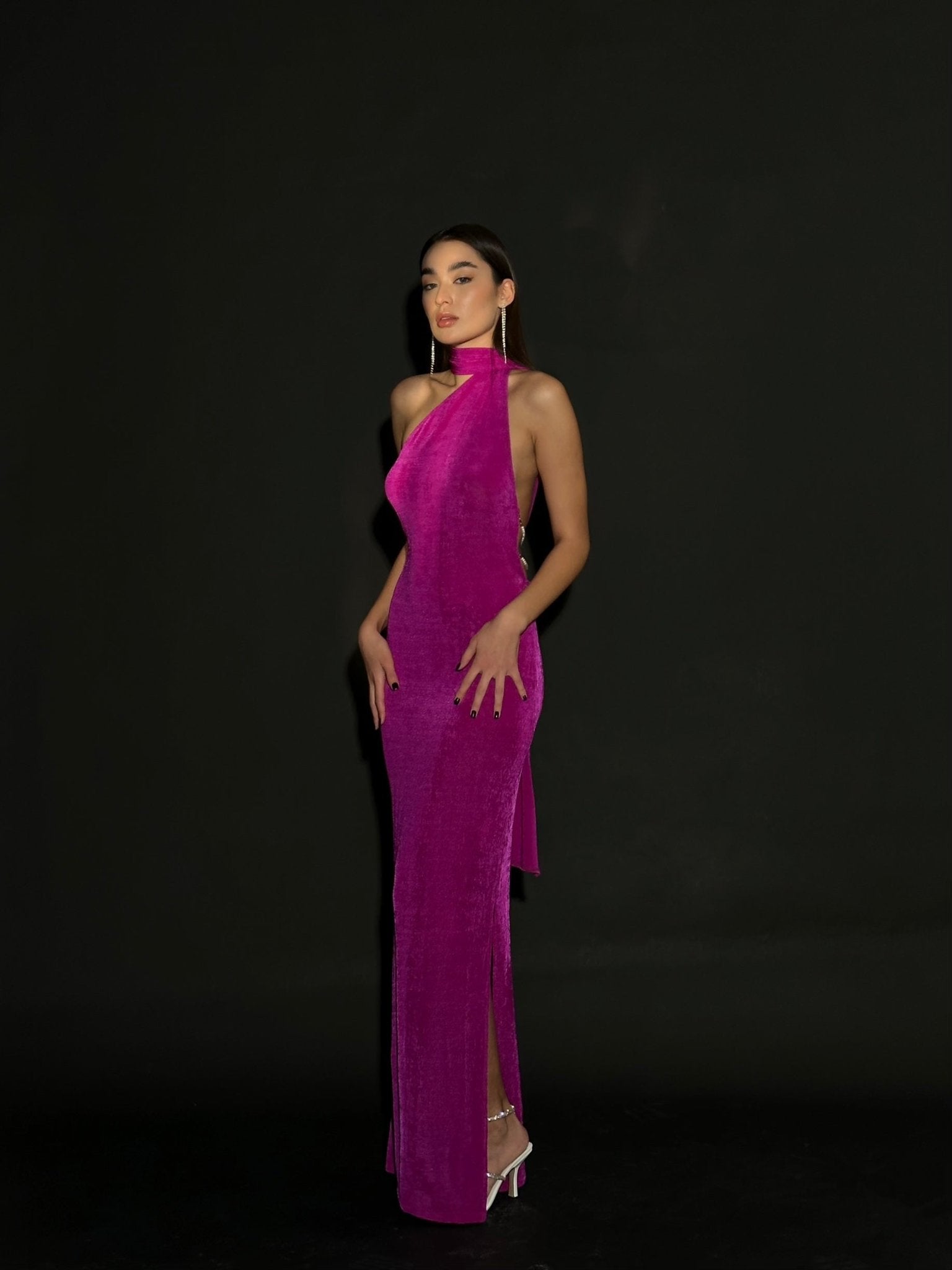 A woman stands confidently against a black background, wearing an Aimée dress in vibrant pink velvet. The gown features a halterneck design with an asymmetrical cut and an open back adorned with pearls. She has long dark hair and is wearing transparent high-heeled shoes.