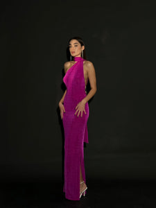 A woman stands confidently against a black background, wearing an Aimée dress in vibrant pink velvet. The gown features a halterneck design with an asymmetrical cut and an open back adorned with pearls. She has long dark hair and is wearing transparent high-heeled shoes.