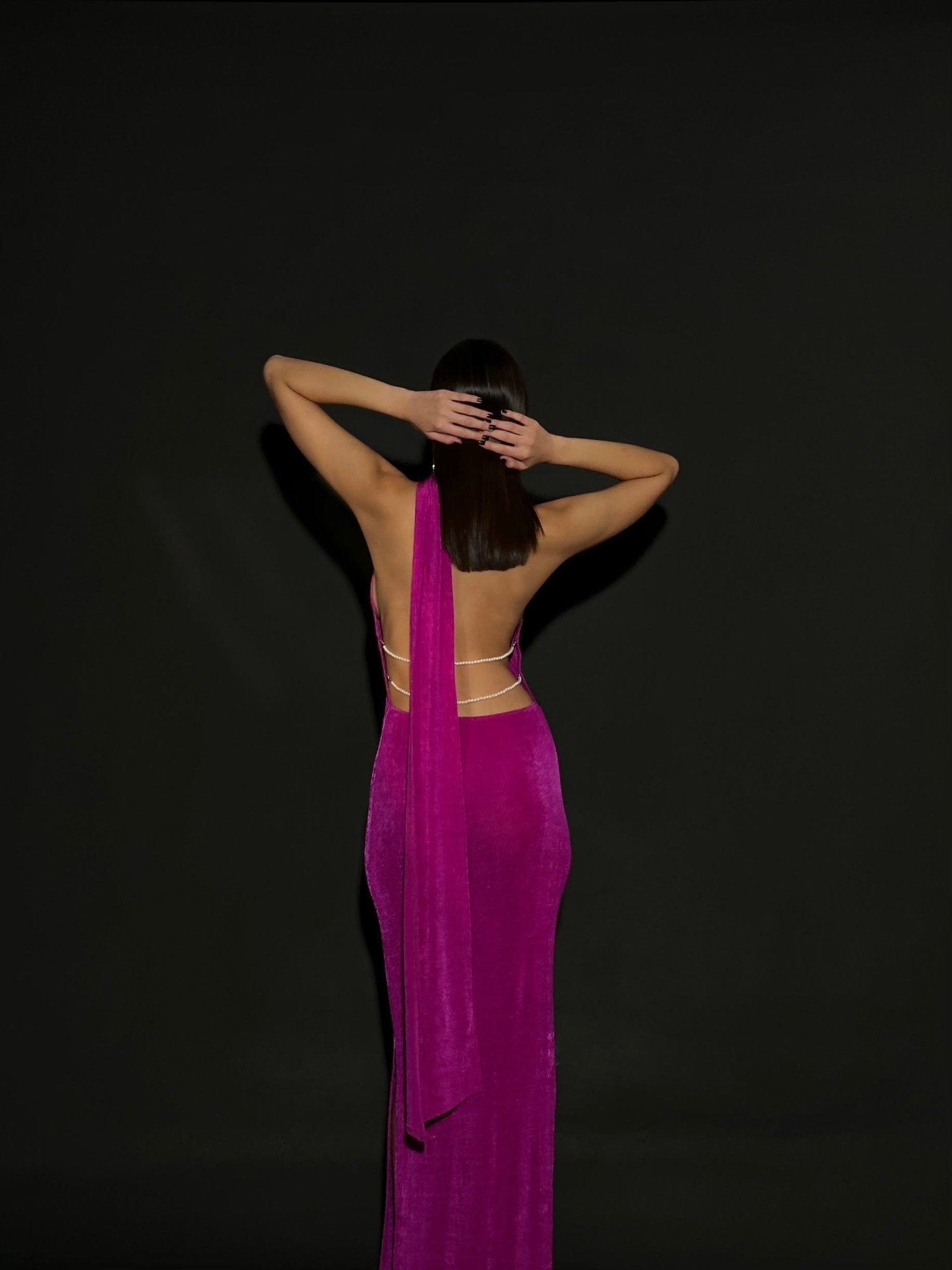 A woman stands with her back to the camera, her hands placed on the back of her head. She is wearing a stunning Aimée dress in vibrant fuchsia, featuring an open back adorned with pearls and thin straps, set against a plain black background.