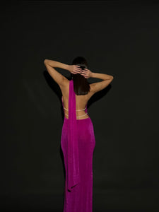 A woman stands with her back to the camera, her hands placed on the back of her head. She is wearing a stunning Aimée dress in vibrant fuchsia, featuring an open back adorned with pearls and thin straps, set against a plain black background.