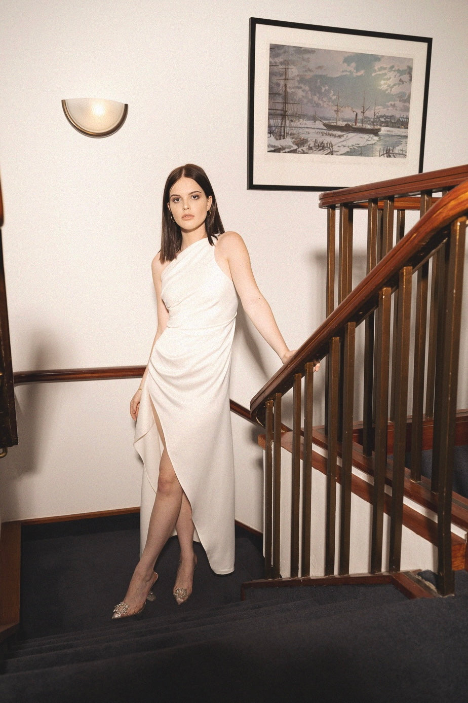 A woman with long dark hair stands on a staircase, wearing the elegant Gianna dress, featuring a one-shoulder design and an asymmetrical hem, paired with sparkling heels. She poses gracefully with one hand on the railing while a framed picture and a wall-mounted light fixture adorn the background.