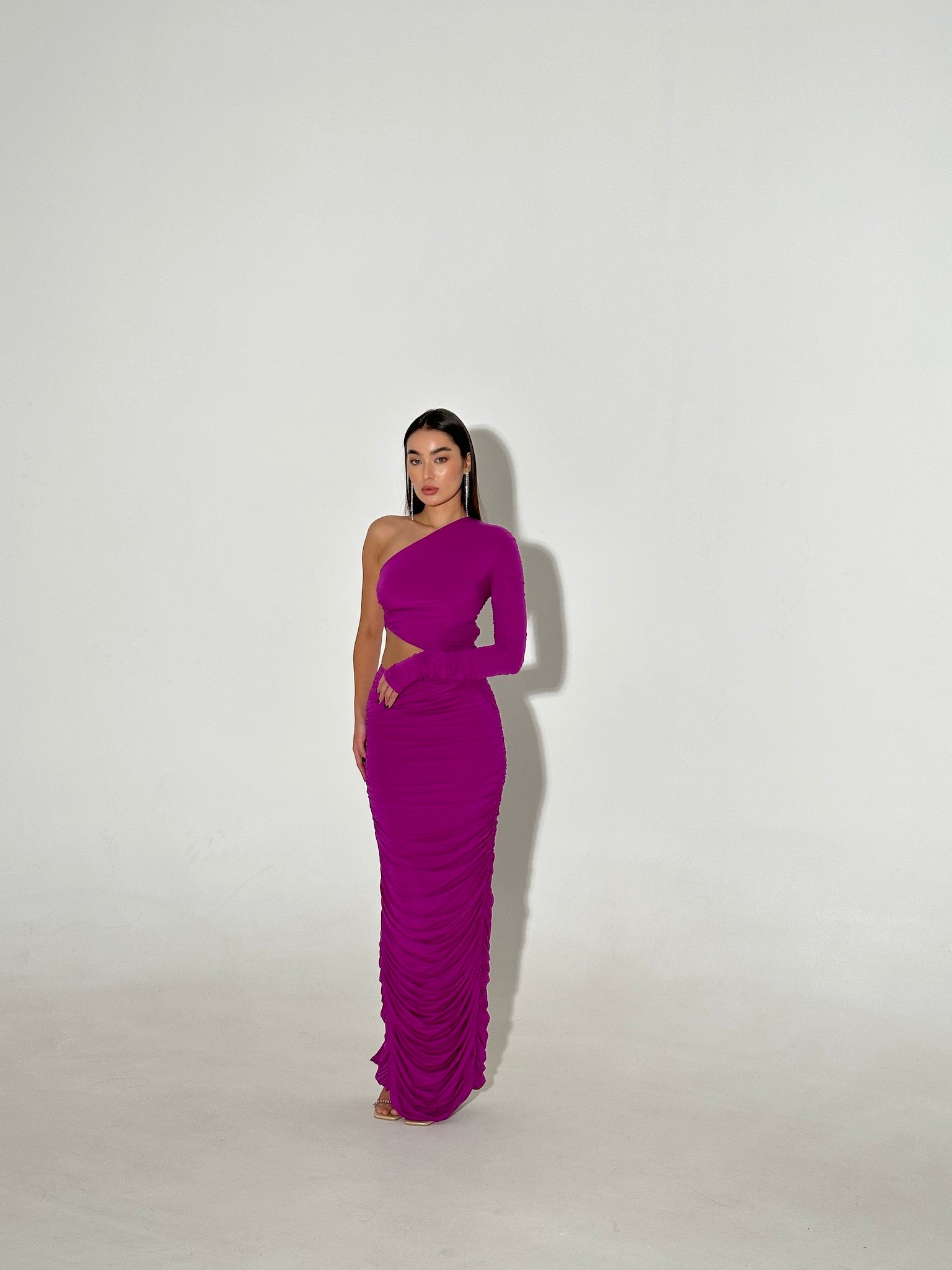 A woman stands against a plain white background, wearing a fitted, vibrant purple Amicia dress that reaches the floor. She has long, straight hair and holds one hand at her hip. The dress features ruched fabric detailing and adjustable straps for added comfort.