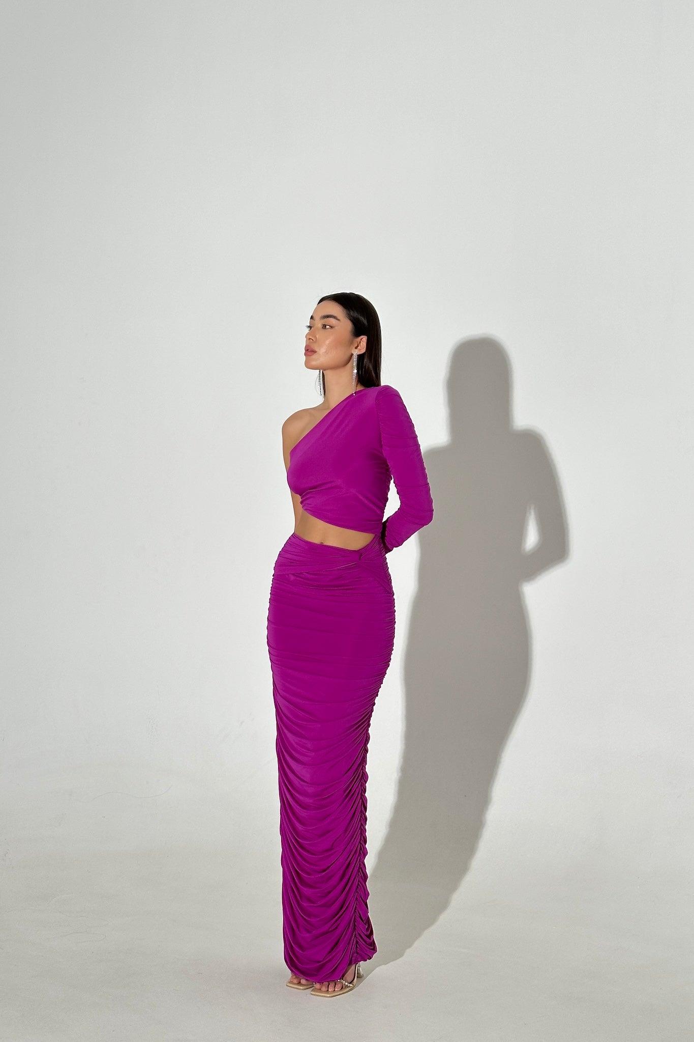 A woman stands against a plain white background, wearing an Amicia dress in vibrant purple, featuring a one-shoulder, long-sleeve crop top design paired with a matching high-waisted, ruched skirt. She has straight dark hair and poses with her hands behind her back, casting a distinct shadow.