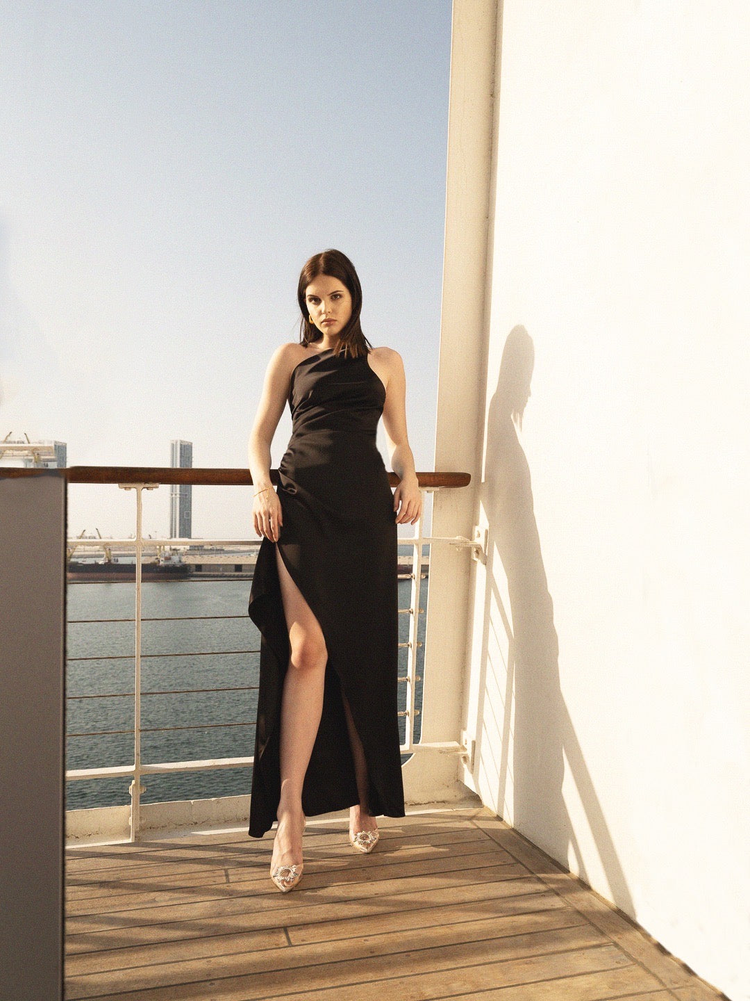 A woman wearing the Gianna dress, featuring a thigh-high slit and an asymmetrical hem in black silky fabric, stands on a balcony overlooking a scenic water view with a cityscape beyond. She leans against the railing as sunlight casts her shadow on the wall behind her, complementing her high heels.