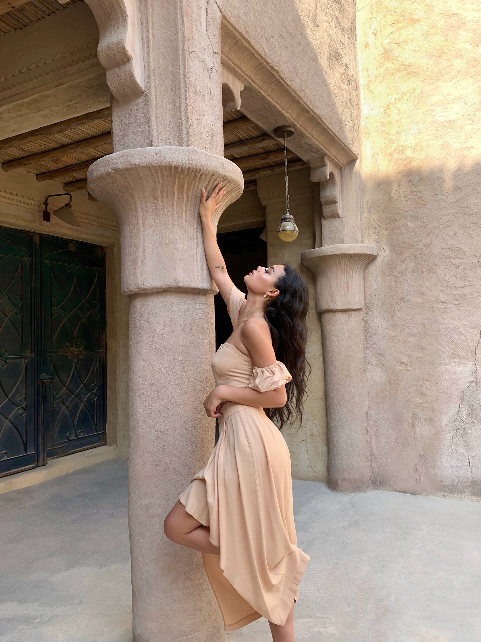 A woman in the Jasmine Set, characterized by its beige hue and linen-like texture, arches her back gracefully against a tall, ornate column. Her arm reaches upward while her long hair cascades down her back. The setting is accented by textured beige walls and a hanging light fixture.