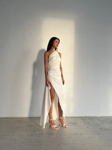 A person stands against a plain background, wearing the Gianna dress, a one-shoulder, silky piece featuring an asymmetrical hem and a high slit, paired with strappy white heels. Light casts a subtle shadow behind them.