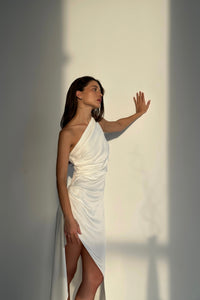 A woman in a Gianna dress, characterized by its one-shoulder white silky fabric and asymmetrical hem with a thigh-high slit, stands indoors against a plain wall lit by sunlight. Her left hand rests gently on the wall while she gazes thoughtfully into the distance, revealing part of her leg through the stylish slit.