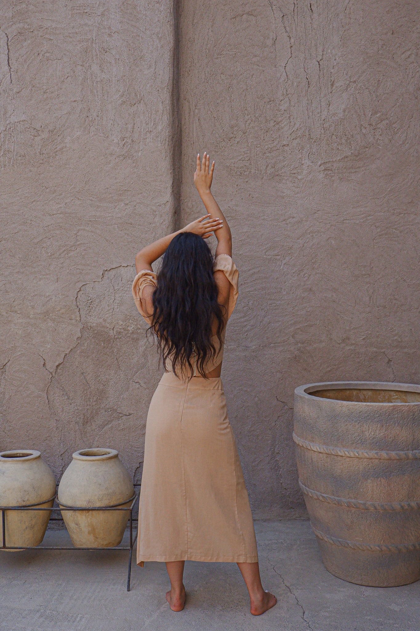 A woman with long hair stands barefoot against a textured beige wall, facing away with her arms raised above her head. She is wearing the Jasmine Set, which includes a linen mix high-low skirt and a matching crop top. Nearby on the ground are two large pots.