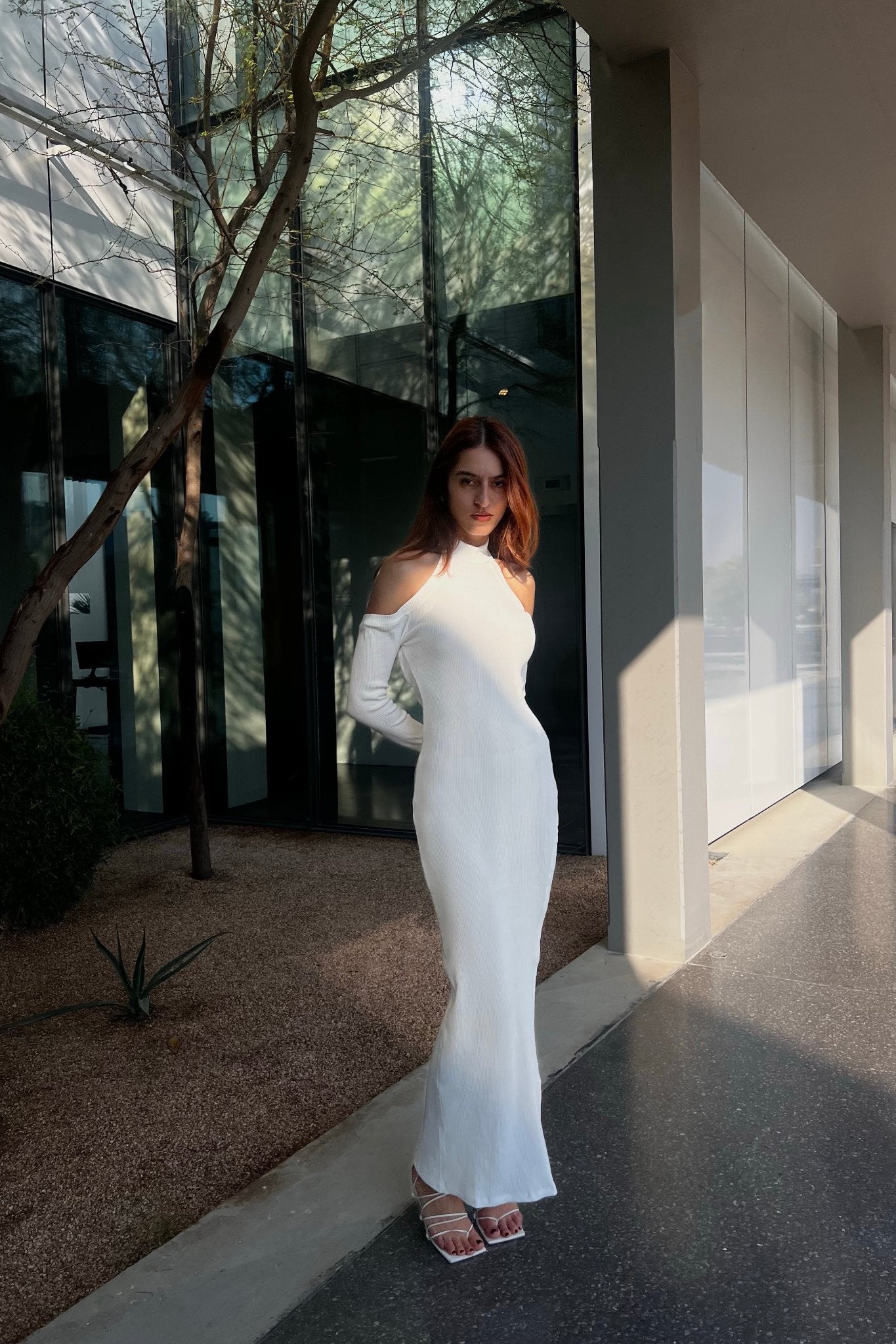 A woman stands confidently in a minimalist setting, wearing the Alara dress. The dress, crafted from breathable cotton fabric, features stylish cut-out shoulders. She pairs it with white strappy heels and her long brown hair flows freely as she stands beside a slender-branched tree, casting intricate shadows on the glass wall behind her.