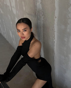 A woman with dark hair pulled back, wearing an Alara dress featuring long, detached sleeves crafted from breathable cotton, leans against a textured, partially painted concrete wall. She gazes at the camera with a serious expression in a dimly lit setting.