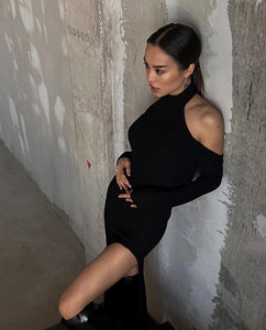 A woman with straight, dark hair leans against a textured, unfinished wall. She is wearing the Alara dress made from breathable cotton fabric, which features cut-out shoulders and a high slit, paired with black boots. Her gaze is directed downward, and her hand rests on her hip.