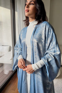 A person with long, dark hair stands near a window, gazing outside. They are wearing the Alara dress—a chic white turtleneck sweater featuring cut-out shoulders—and have a shimmering blue robe draped over it. The room has a modern design with a wooden floor and white furniture that is beautifully illuminated by natural light.